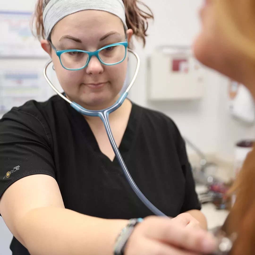 Healthcare professional wearing teal glasses and a headband using a stethoscope to listen to a patient's heartbeat at Highland Health Providers.
